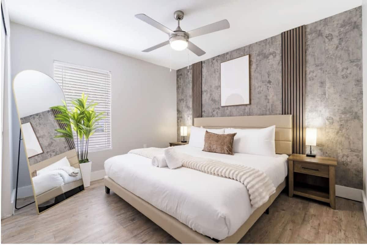 Un dormitorio moderno con cama, mesitas de noche, ventilador de techo, planta en maceta y un amplio ventanal con persianas.