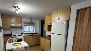 A compact kitchen with wooden cabinets, a white refrigerator, a stove, and a dining table with a chair. - Charming, cozy studio 7 minutes from Ottawa (Gatineau)