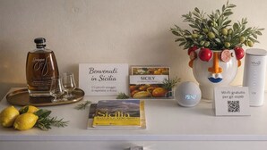 Et hotelværelse med en Grappa-flaske, en velkomstskilt, en bog med titlen 'Sicilia', en dekorativ vase med frugt, et ur og et Wi-Fi-skilt.