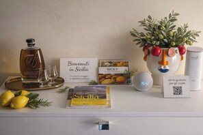 Une chambre d’hôtel avec une bouteille de grappa, un panneau d’accueil, un livre intitulé « Sicile », un vase décoratif rempli de fruits, une horloge et un panneau indiquant la présence d’une connexion Wi-Fi.