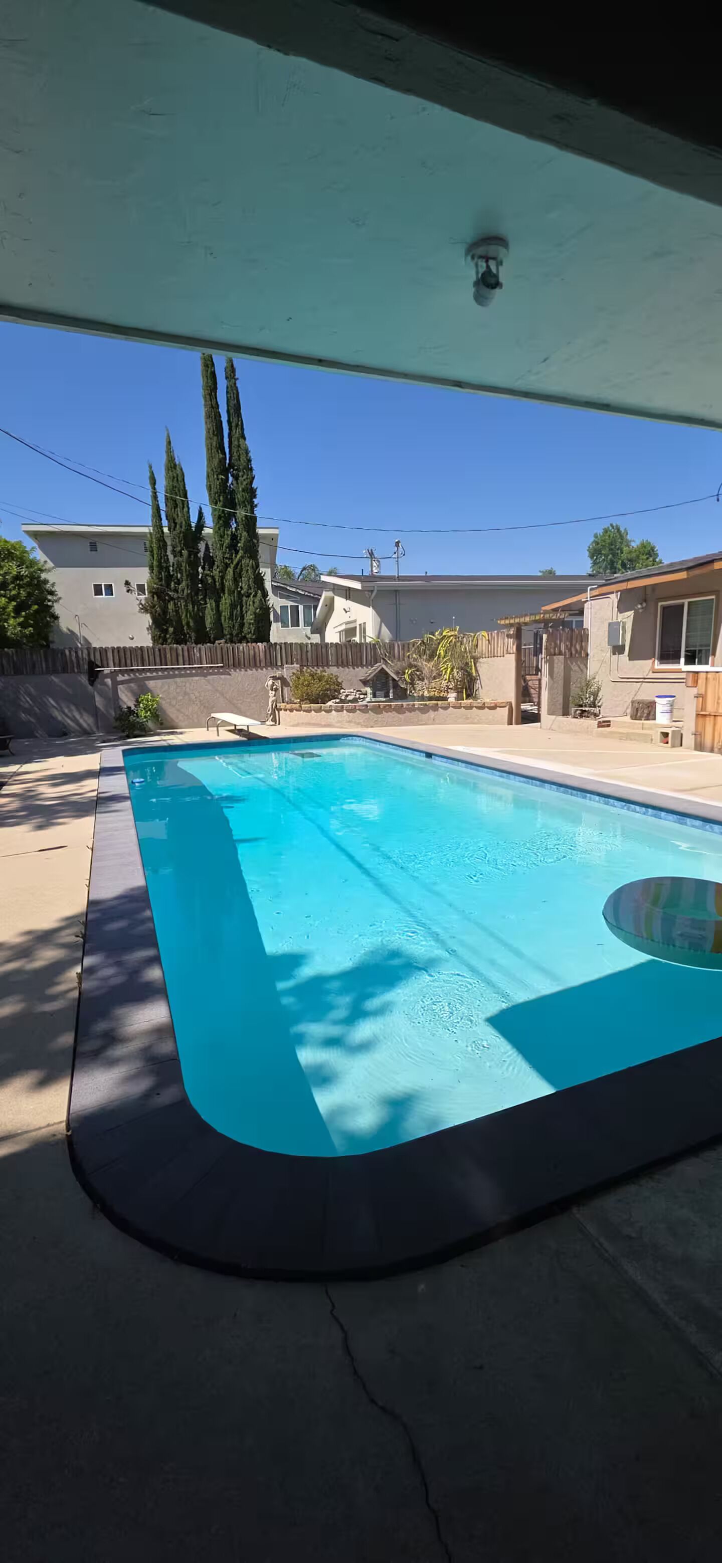 Ein Pool mit Holzterrasse, überdachtem Bereich und Blick auf Häuser im Hintergrund.