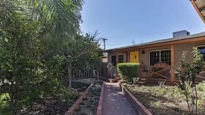 Un sendero conduce a un edificio con puerta amarilla, rodeado de follaje y un borde de ladrillo.