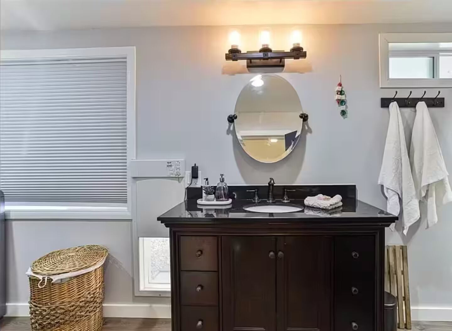Un baño con tocador oscuro, un espejo redondo, una lámpara, un perchero y una cesta de mimbre.