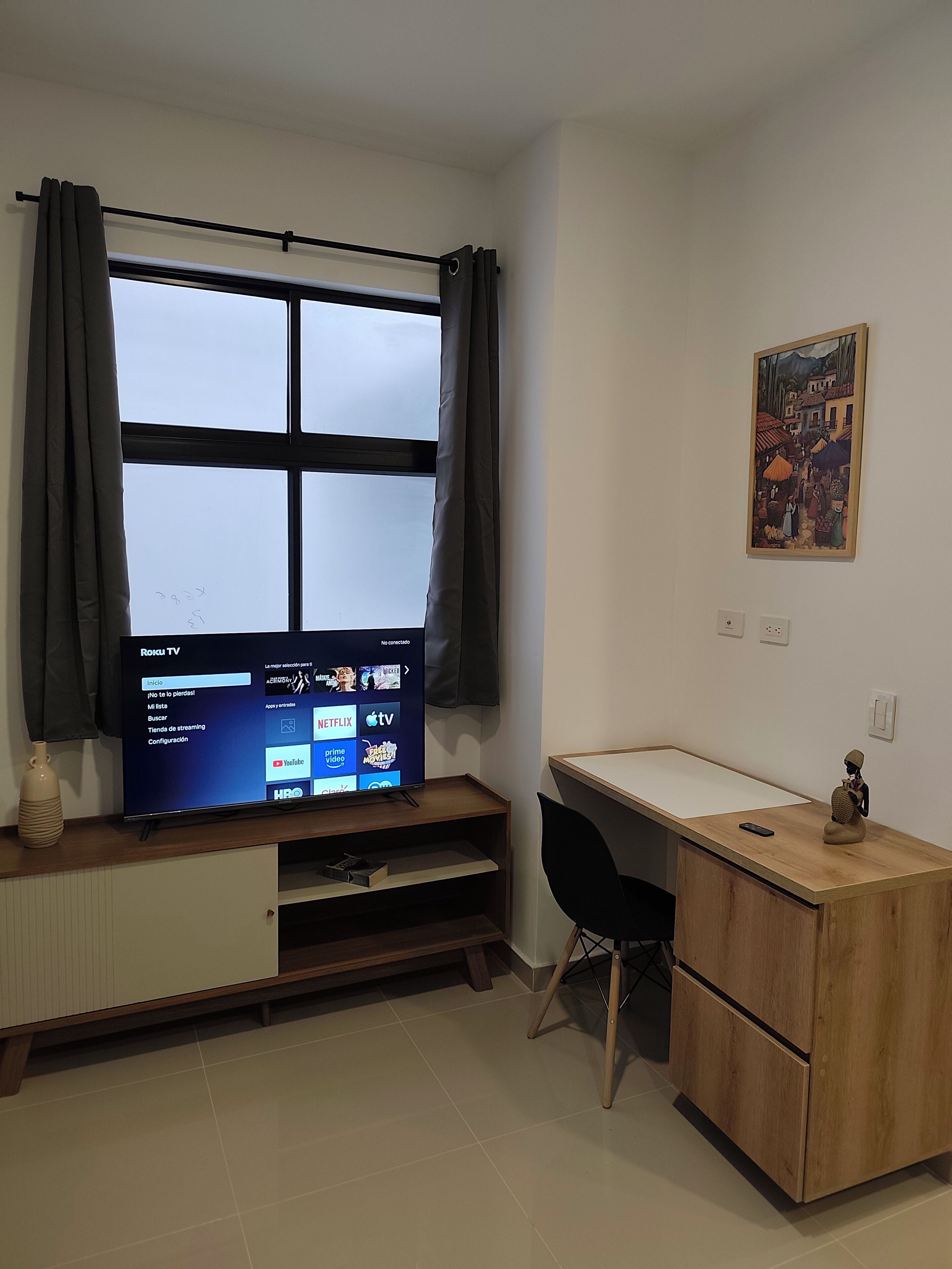 A room with a TV on a wooden stand, a desk with a chair, a small statue, and a painting on the wall.