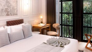 Habitación de hotel con una cama grande, un escritorio, una silla y vistas a zonas verdes a través de amplios ventanales.