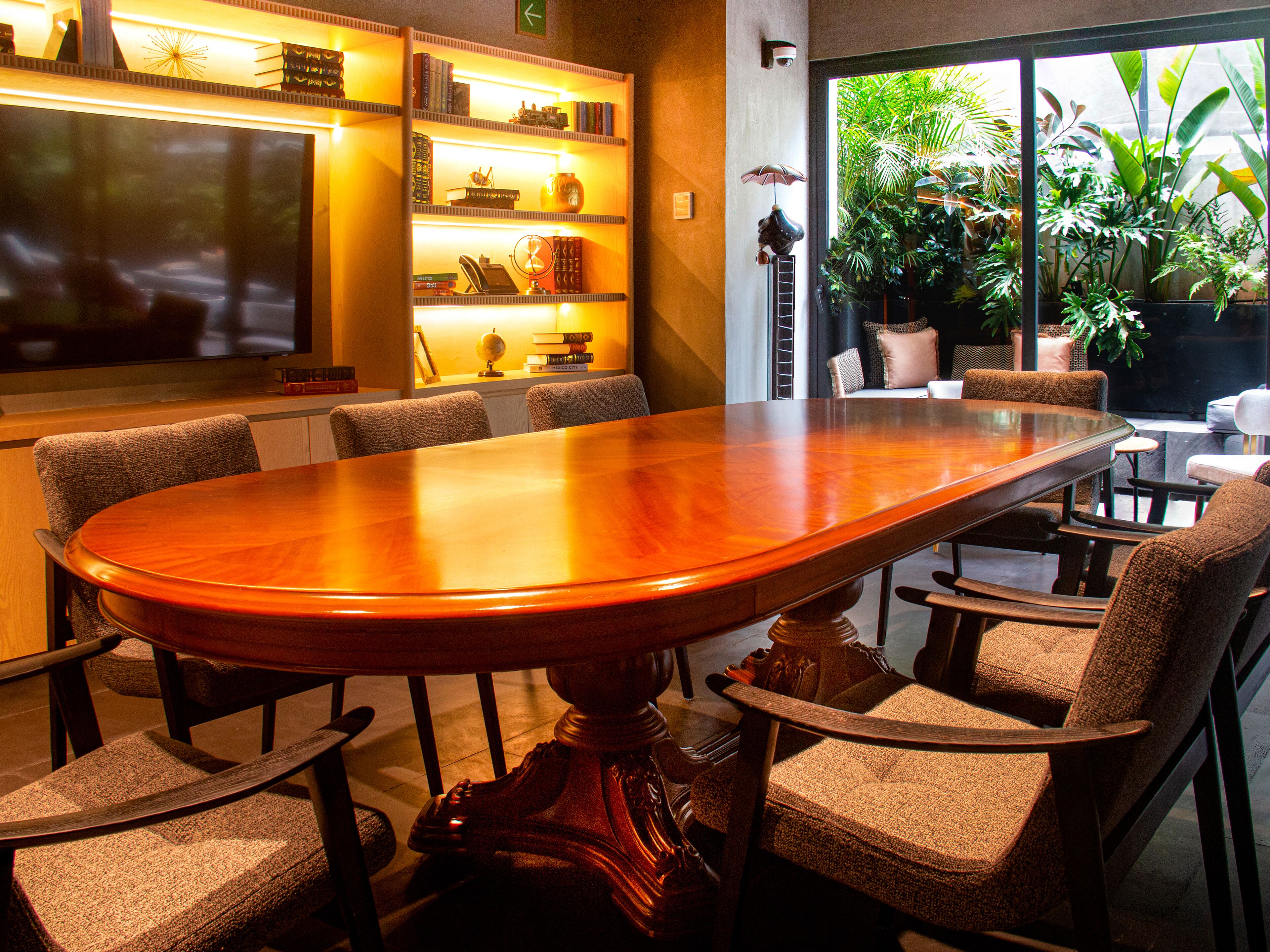 A conference table with chairs, a television, shelves with books and decorative items, and a view of greenery through a window.