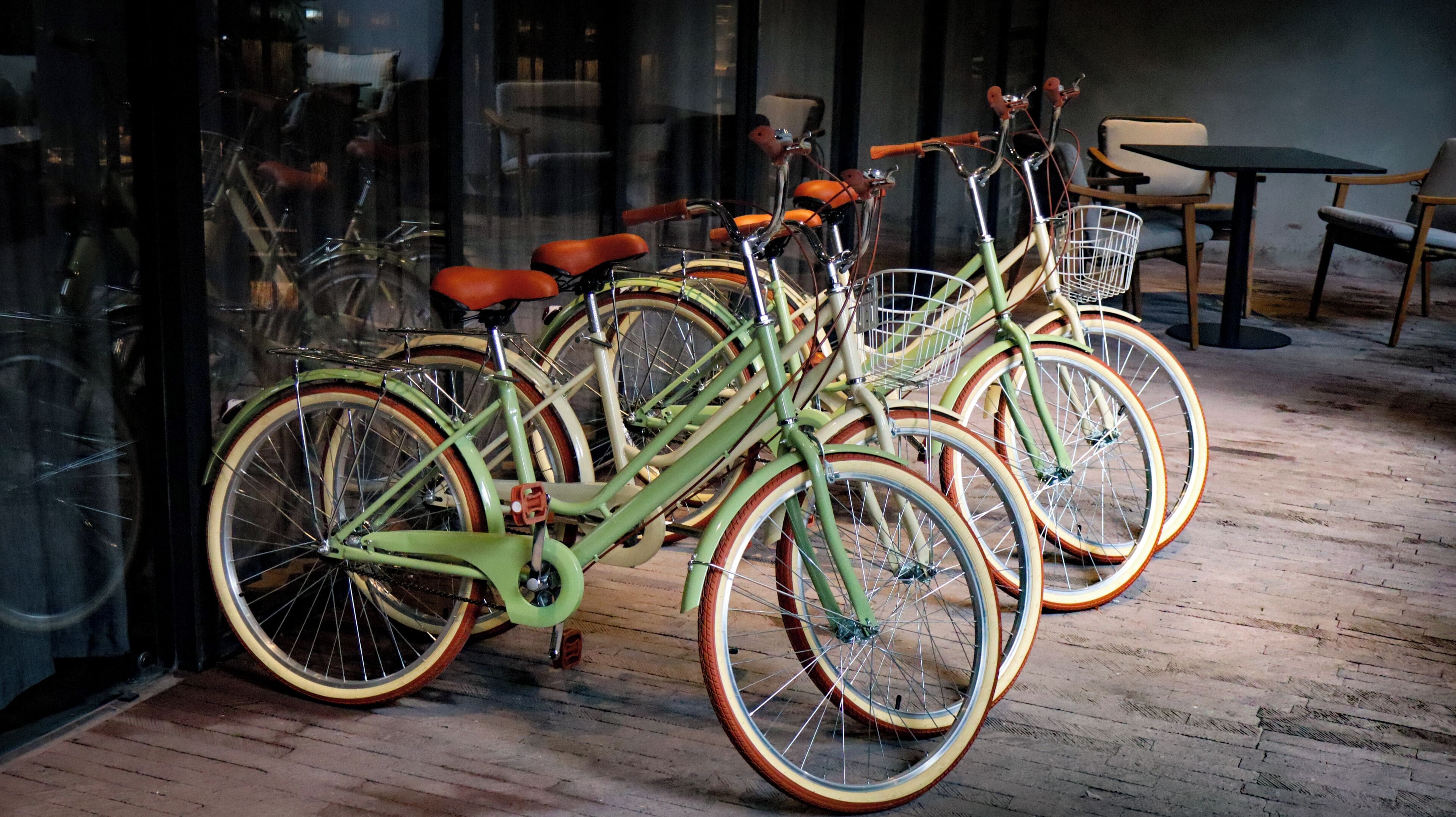 Una hilera de bicicletas antiguas con cestas, suelo de madera y un escaparate de vidrio que refleja muebles del interior.