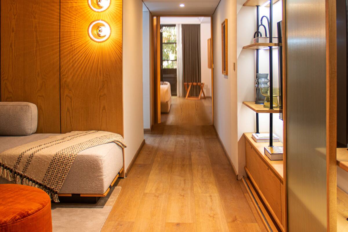 Quarto de hotel com cama, guarda-roupa de madeira, mesa de cabeceira e prateleira com objetos decorativos.