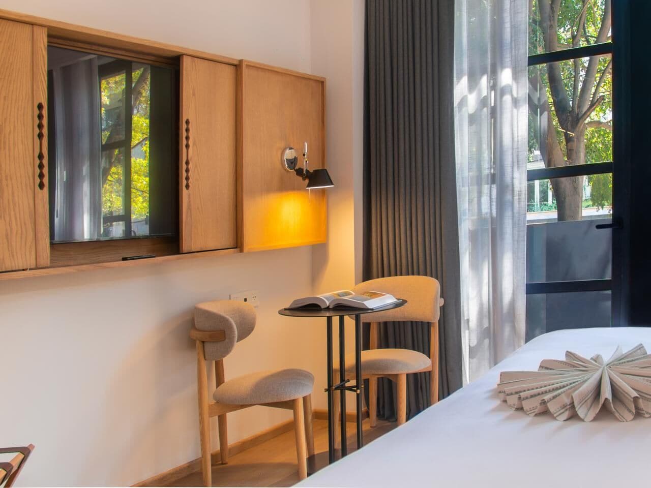 Une chambre d’hôtel avec un lit, une petite table avec des chaises, une lampe et une fenêtre avec des rideaux.
