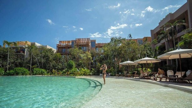 A swimming pool with clear water, surrounded by lounge chairs and umbrellas, and buildings in the background. - Executive Unit in Aldea Zama and Parking (Tulum)