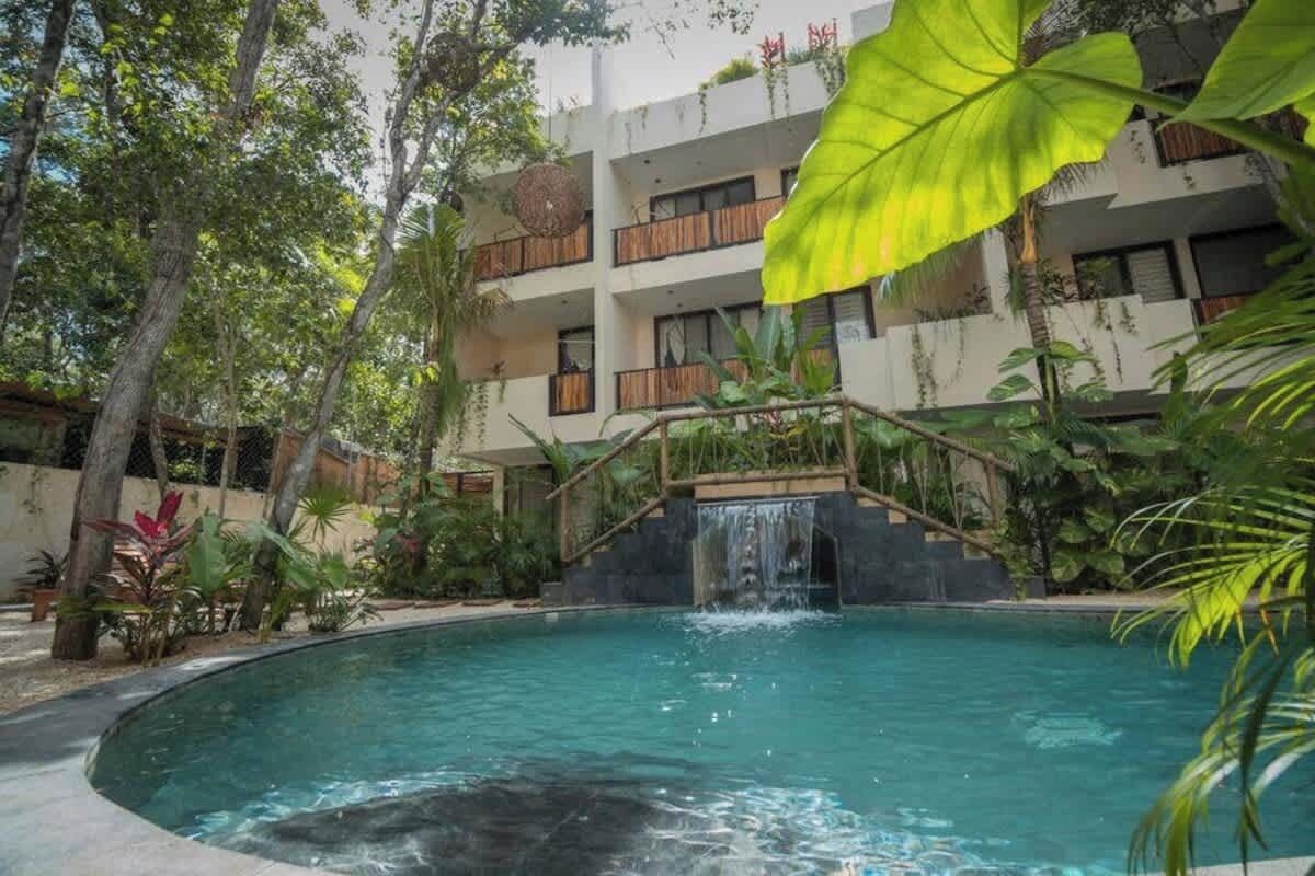 A pool with a waterfall feature, surrounded by lush greenery and a multi-story building with balconies.