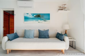 Een witte bank met blauwe kussens, een schilderij van een boom aan het strand, een lamp en een houten vloer.