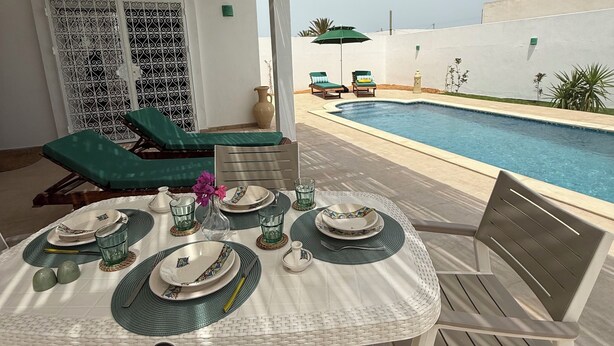 Un espace de restauration extérieur abrité, avec vue sur la piscine, une table dressée avec des assiettes, des verres et des couverts, ainsi que deux chaises longues.
