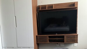Une pièce avec une armoire blanche, un grand téléviseur à écran plat fixé sur un panneau en bois et un pouf gris.