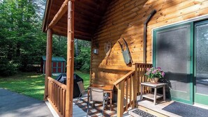 Una baita di legno con un portico, un tavolo, sedie, un barbecue e una porta con una finestra.