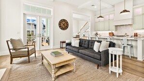 A spacious living room with a sofa, coffee table, and a chair. A kitchen area with a countertop and hanging lights is visible in the background. - Tortuga House 105 SOU (Port Aransas)