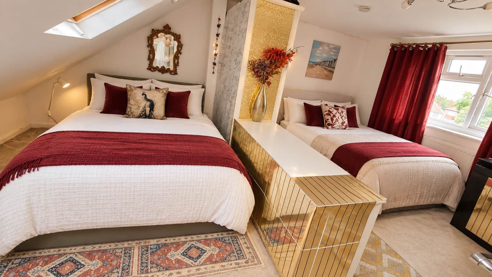 A bedroom with two beds, a decorative mirror, a vase with flowers, and a view of the outdoors.