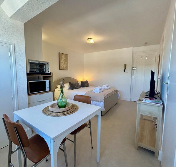 A compact room with a bed, a small table with chairs, a kitchenette, and a television on a stand.