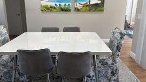Un'area pranzo con un tavolo bianco, quattro sedie grigie e due quadri a tema spiaggia.