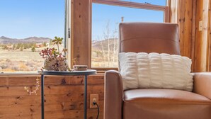A room with a brown leather chair, a white fluffy pillow, a small table with a plant, and a window with a view of a desert landscape. - Aspen Grove Cabin - Sauna, 3 Decks, Mountain Views, ATV Access (Grand Lake)