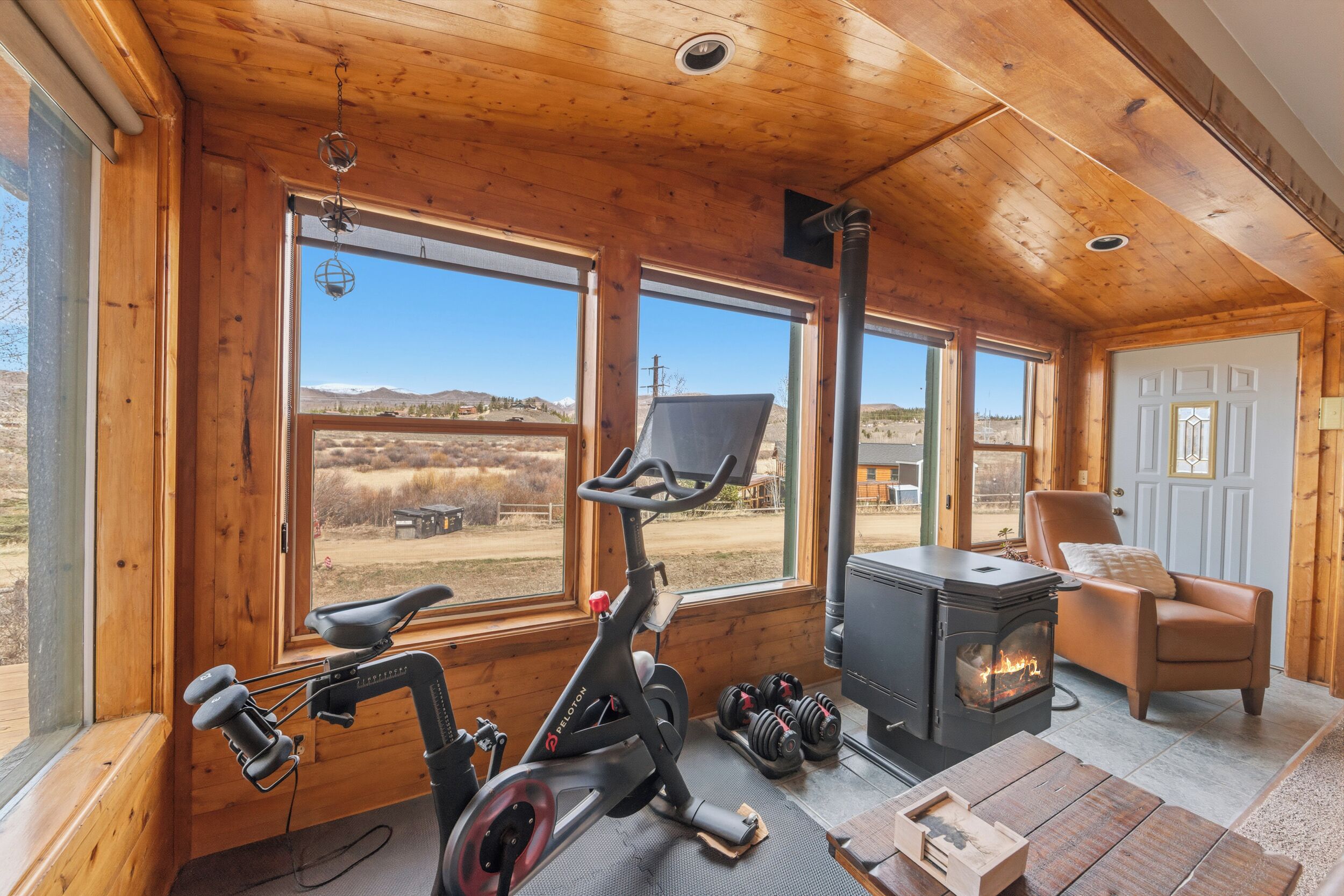 A wooden cabin interior with a stationary bike, dumbbells, a fireplace, and large windows.