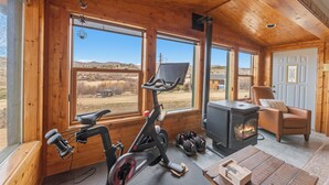 A wooden cabin interior with a stationary bike, dumbbells, a fireplace, and large windows. - Aspen Grove Cabin - Sauna, 3 Decks, Mountain Views, ATV Access (Grand Lake)