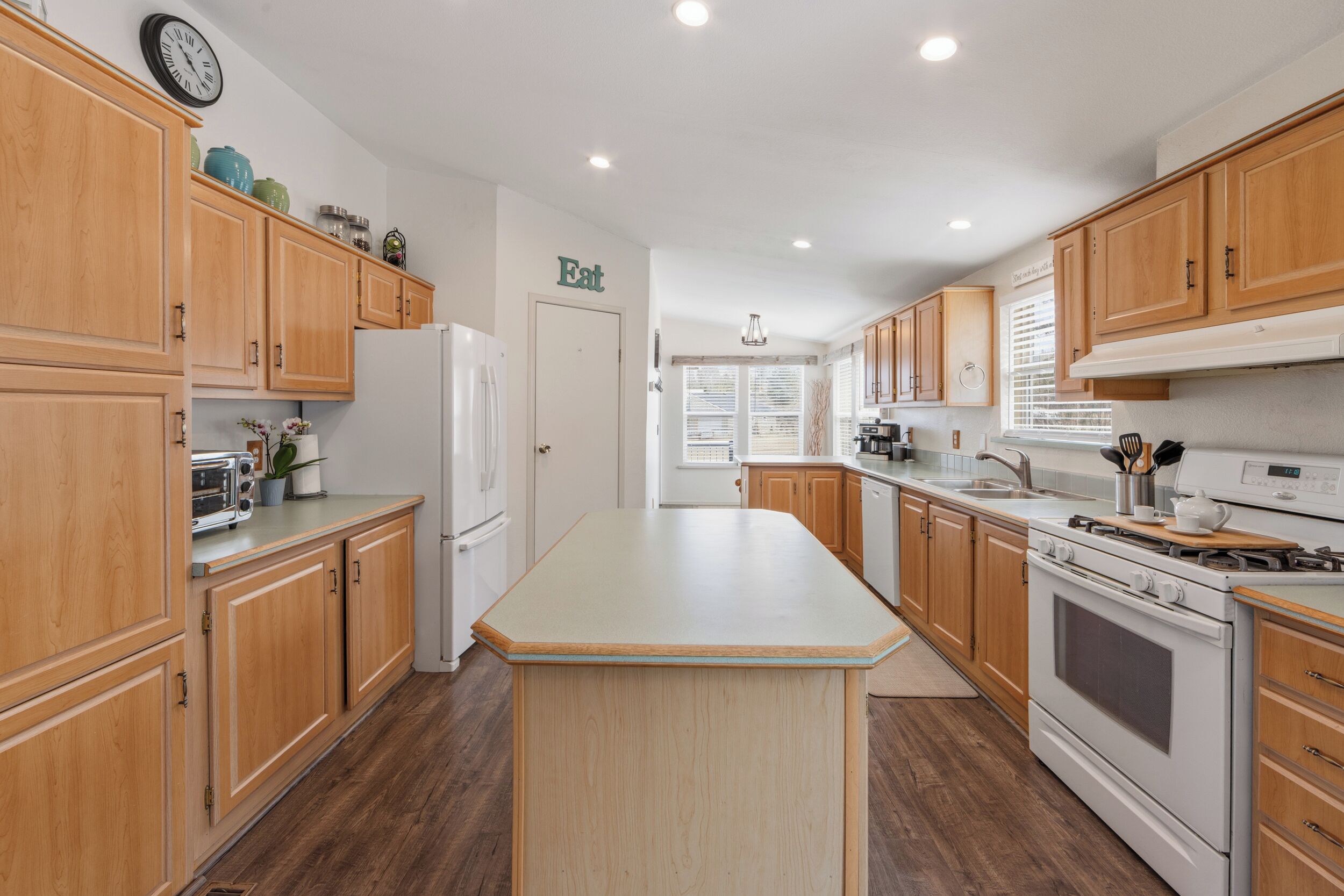 A kitchen with wooden cabinets, a central island, a refrigerator, an oven, and a clock on the wall.