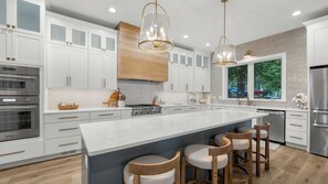 Une cuisine moderne avec des meubles blancs, un grand îlot central doté de tabourets, des appareils en acier inoxydable et une fenêtre offrant une vue sur l’extérieur.