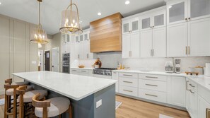 Uma cozinha moderna com armários brancos, uma ilha central com banquetas, eletrodomésticos de aço inoxidável e iluminação pendente.