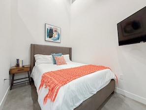 Ein Schlafzimmer mit einem Bett, einem Nachttisch, einem an der Wand montierten Fernseher und einem gerahmten Bild über dem Bett.