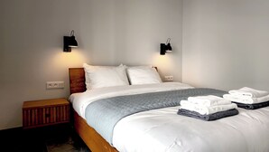 Une chambre d’hôtel avec un lit en bois, du linge de lit blanc, deux lampes fixées au mur, une table de chevet et une serviette pliée posée sur le lit.
