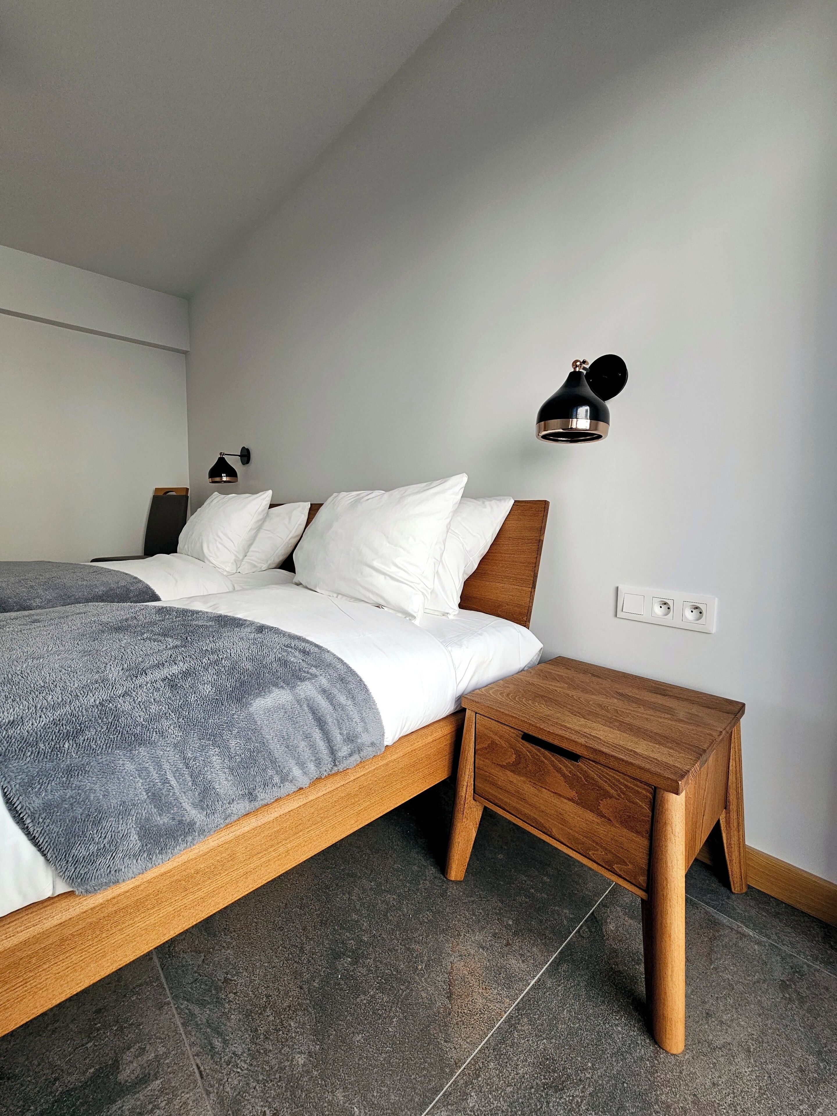 A modern bedroom with a wooden bed, a bedside table, a wall-mounted lamp, and a power outlet.