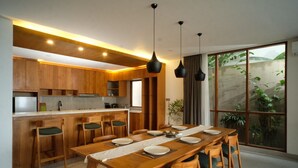 A modern kitchen with wooden cabinets, a dining table set for six, and a large sliding glass door to a garden. - THE LEDGE ULUWATU (Pecatu)