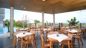 An outdoor dining area with wooden tables and chairs, a pool, and a view of the landscape. - THE LEDGE ULUWATU (Pecatu)