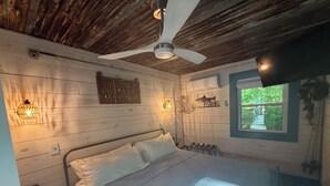 Um quarto rústico tipo cabana com cama, ventoinha no teto, luminárias fixadas na parede e uma janela com vista para o verde.