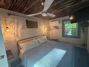 Une chambre de style cabane en bois, avec un lit, un ventilateur de plafond, des lampes fixées au mur et une fenêtre donnant sur un paysage verdoyant.
