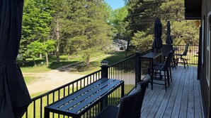 Um deck de madeira com mobiliário externo, vista para uma floresta e céu claro.