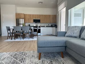 Una cocina moderna con armarios de madera, un comedor con mesa y sillas, y un sofá gris con cojines estampados.