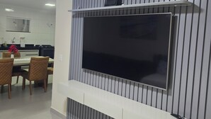 A modern living room with a large flat-screen TV mounted on a wall with vertical paneling, a dining area with a table and chairs, and a kitchen in the background. - Luxury apartment 200 meters from the beach. (Porto Seguro)