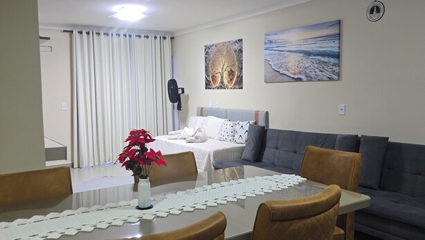 A dining area with a table, chairs, a vase with flowers, a bed with a white blanket, a gray sofa, and two wall art pieces. - Luxury apartment 200 meters from the beach. (Porto Seguro)