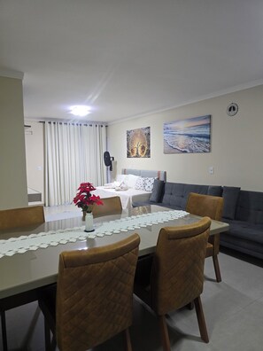 A dining area with a table, chairs, a vase with flowers, a bed with a white blanket, a gray sofa, and two wall art pieces. - Luxury apartment located 200 meters from the beach. (Porto Seguro)