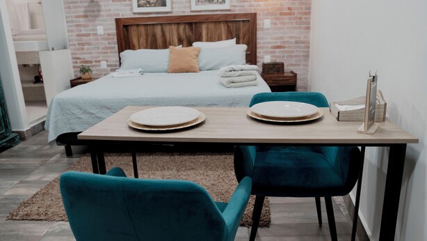 Une chambre d’hôtel avec un lit, une table en bois avec des assiettes, des chaises de couleur turquoise et un mur en briques.