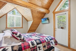 A bedroom with a sloped ceiling, a bed with a colorful quilt, a TV on the wall, and a door with a view of trees.