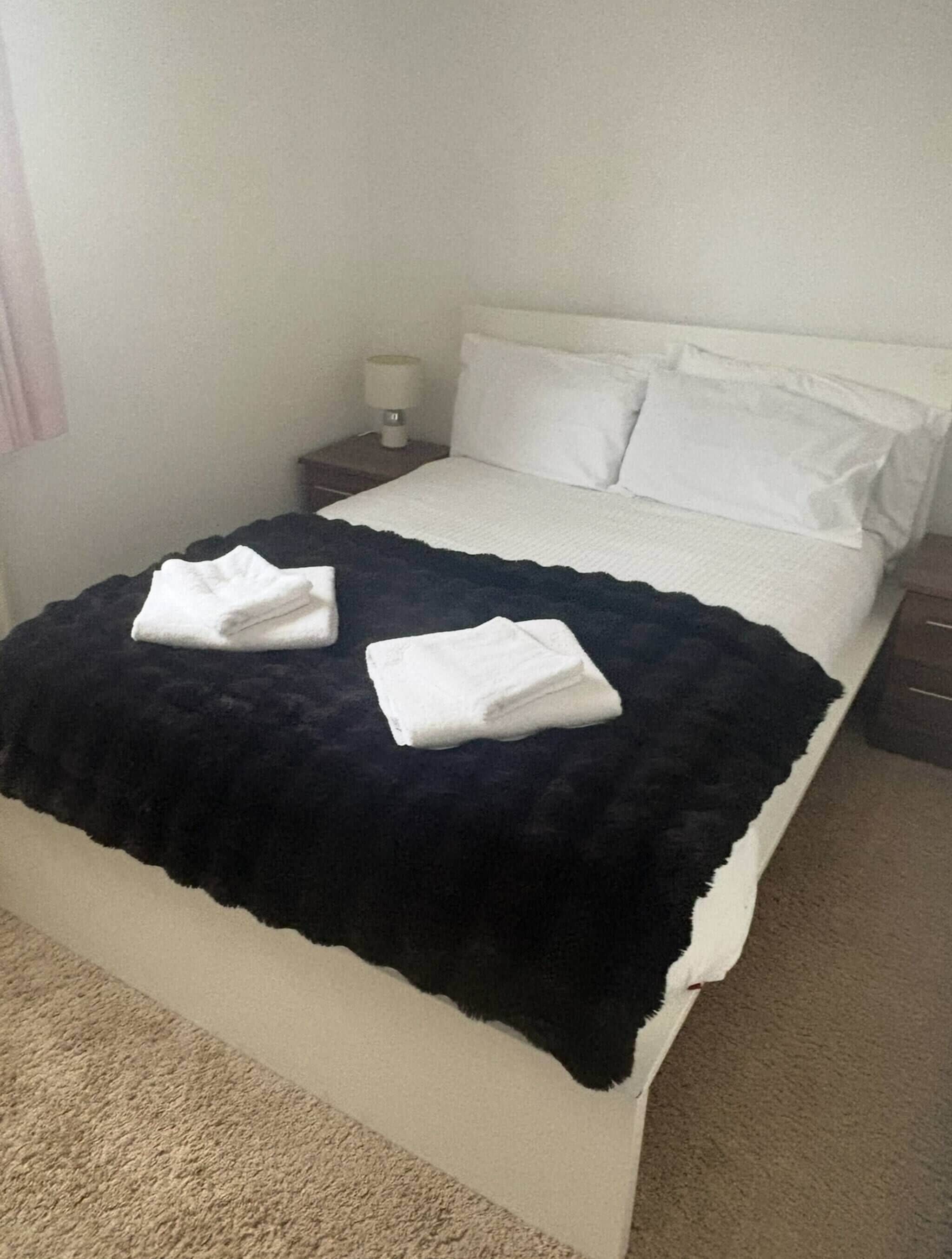Een bed met een zwart deken en twee opgevouwen witte handdoeken, een bedlamp en een nachtkastje.