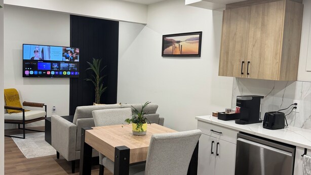 Una cocina moderna con lavavajillas incorporado, un comedor con mesa de madera y sillas grises, un televisor de pantalla plana montado en la pared y una planta en maceta sobre la mesa.