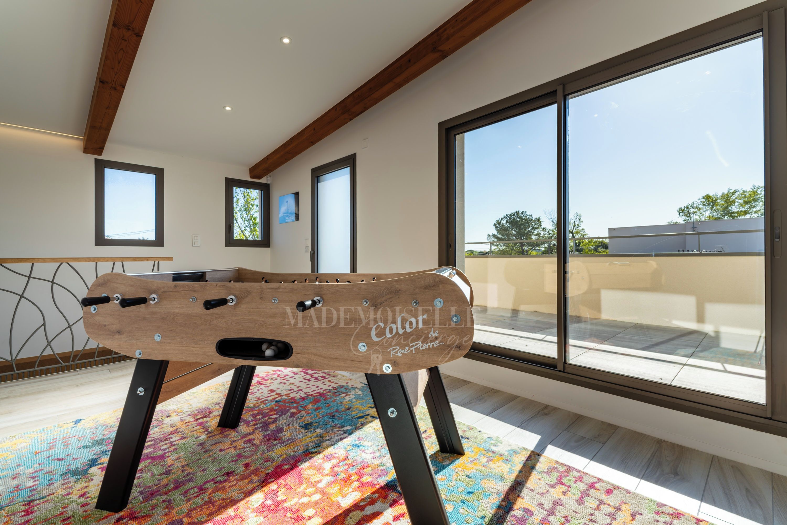 Een kamer met een houten tafelvoetbaltafel, grote ramen en een kleurrijk vloerkleed.