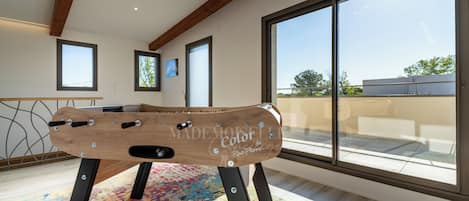 Een kamer met een houten tafelvoetbaltafel, grote ramen en een kleurrijk vloerkleed.