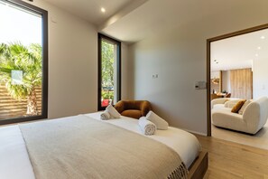 Ein modernes Hotelzimmer mit einem großen Bett, einer Sitzecke mit einer Couch und Blick auf Palmen durch ein Fenster.