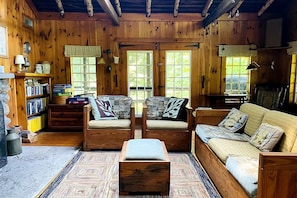 A cozy cabin interior with wooden walls, a sofa, two armchairs, a small table, and a bookshelf.