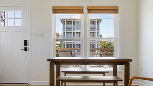 Una habitación con una mesa de madera y un banco, una puerta blanca y ventanas amplias con vistas a un edificio de varios pisos.